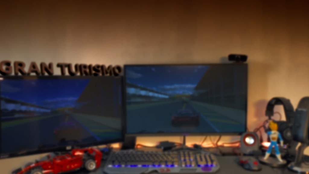 Mesa com dois monitores exibindo Gran Turismo, teclado e volante de simulação